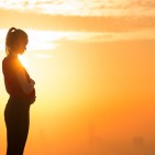 Pregnant woman standing alone holding her belly during sunset. Maternity and motherhood care.