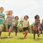 Happy kids, playing and running with grass field in nature for fun, playful day or sunshine at park. Diversity or group of excited children or youth enjoying sunny outdoor holiday or weekend together