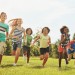 Happy kids, playing and running with grass field in nature for fun, playful day or sunshine at park. Diversity or group of excited children or youth enjoying sunny outdoor holiday or weekend together